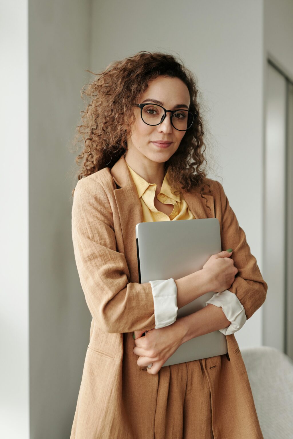 A confident woman in formal attire holding a laptop, standing in an office environment and looking at the camera.